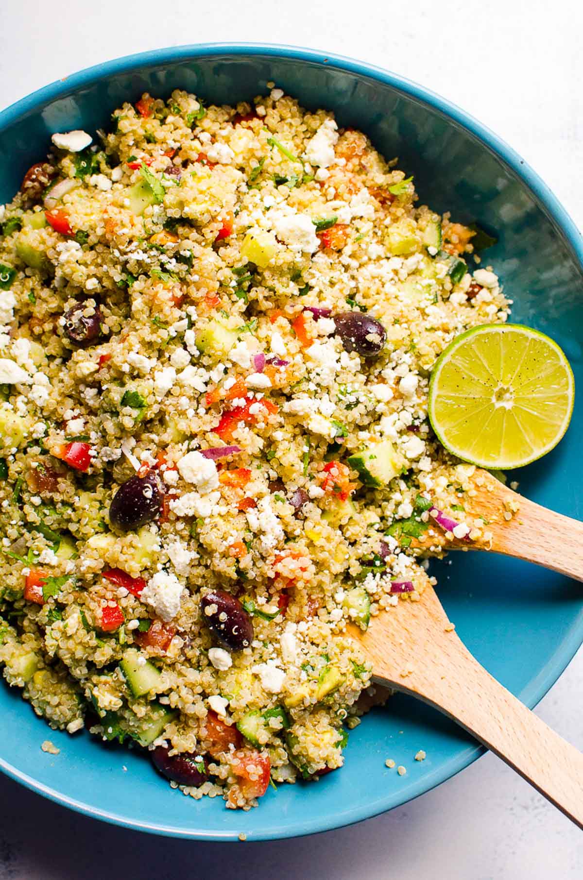 Mediterranean quinoa salad in blue serving bowl with wood tongs and lime.