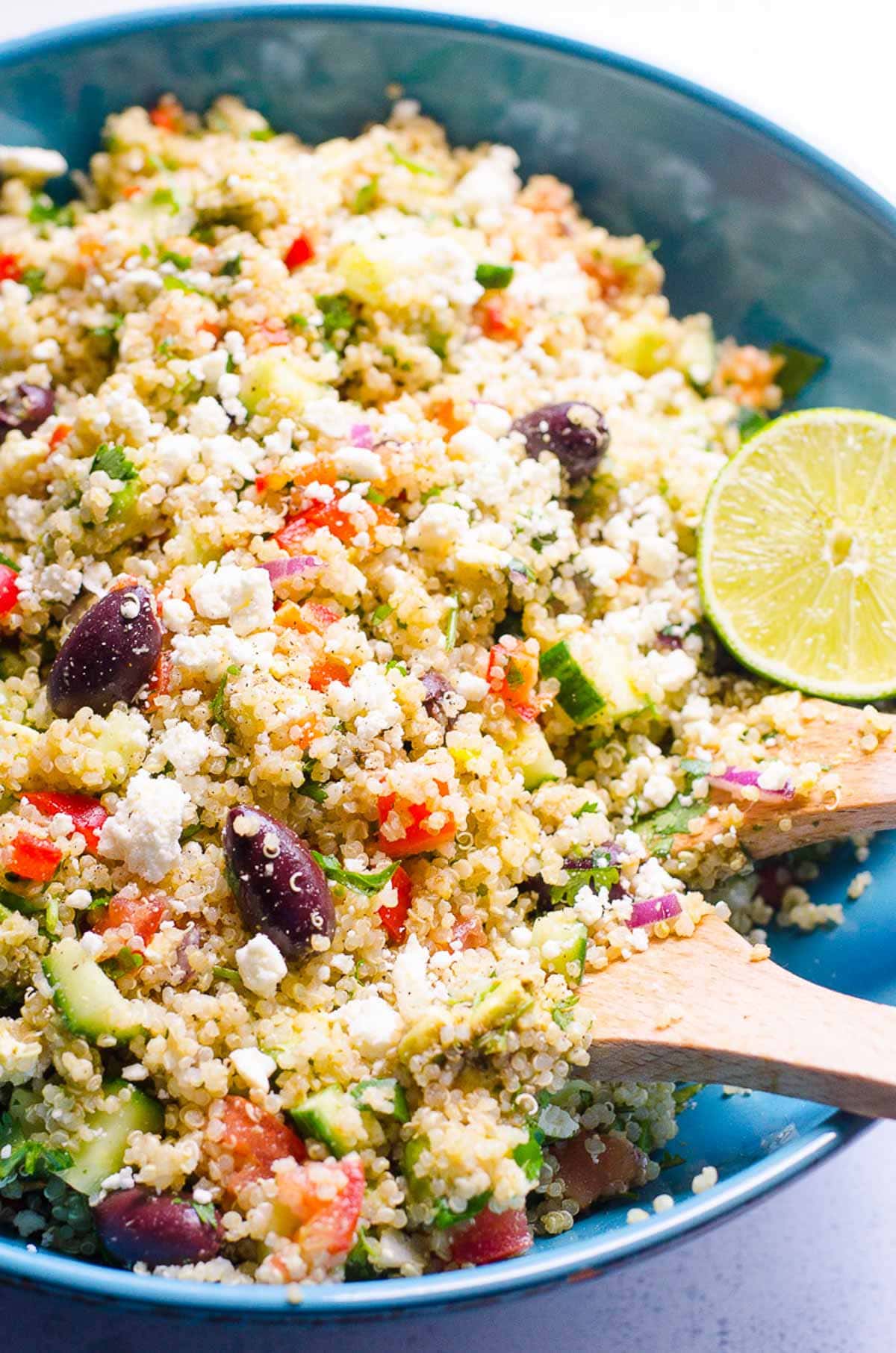 Mediterranean quinoa salad with feta, lime and wooden serving spoons in blue bowl.