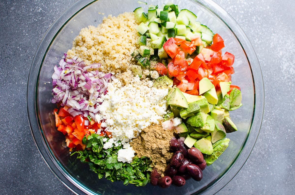 Chopped tomatoes, cucumbers, avocado, feta cheese, red onions and olives in a bowl with quinoa and seasonings.
