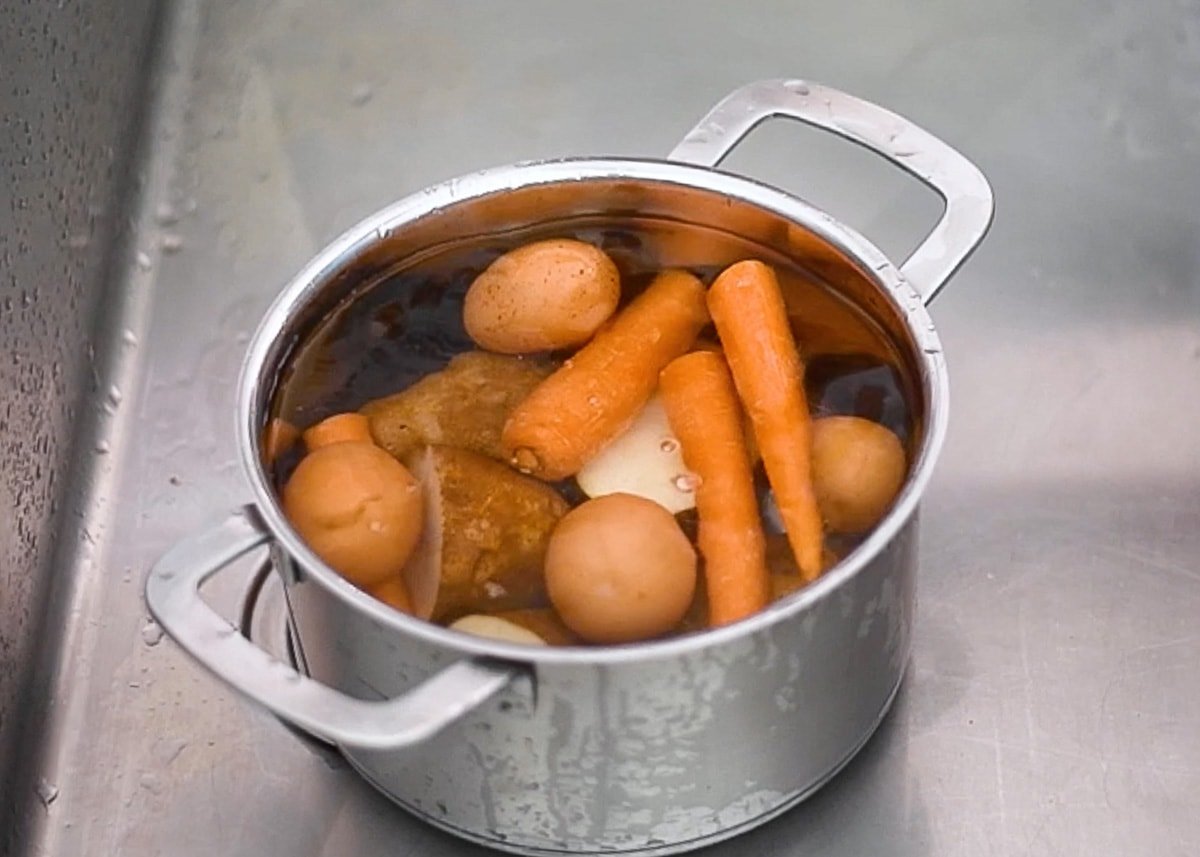 A pot with potatoes, carrots, eggs and cold water in the sink.