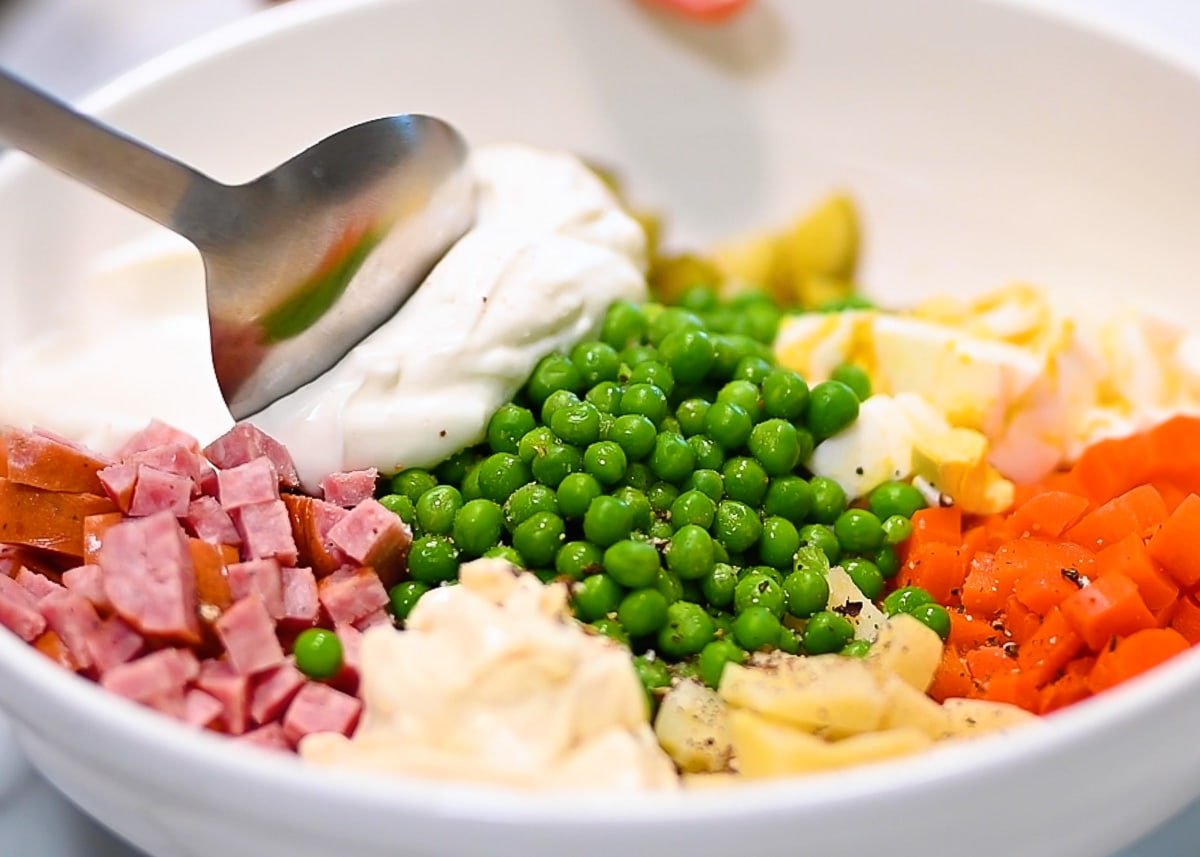 A spoon spreading yogurt and mayo over a bowl with diced sausage, carrots, eggs, potatoes, pickles and peas.