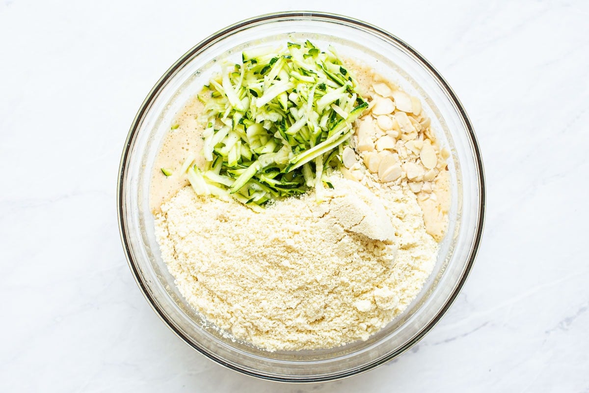 Almond flour, shredded zucchini and walnuts added to glass bowl with liquid ingredients. 