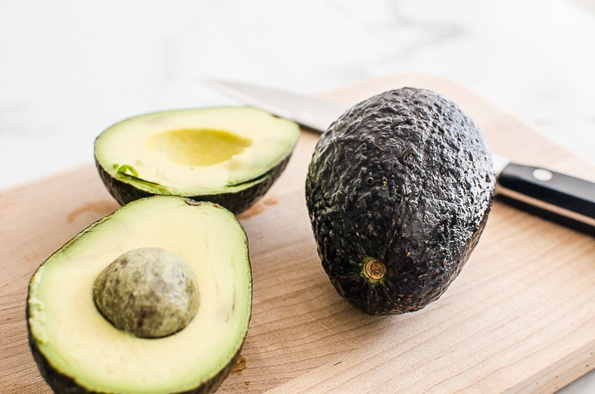 Two avocados on a cutting board with one cut in half.