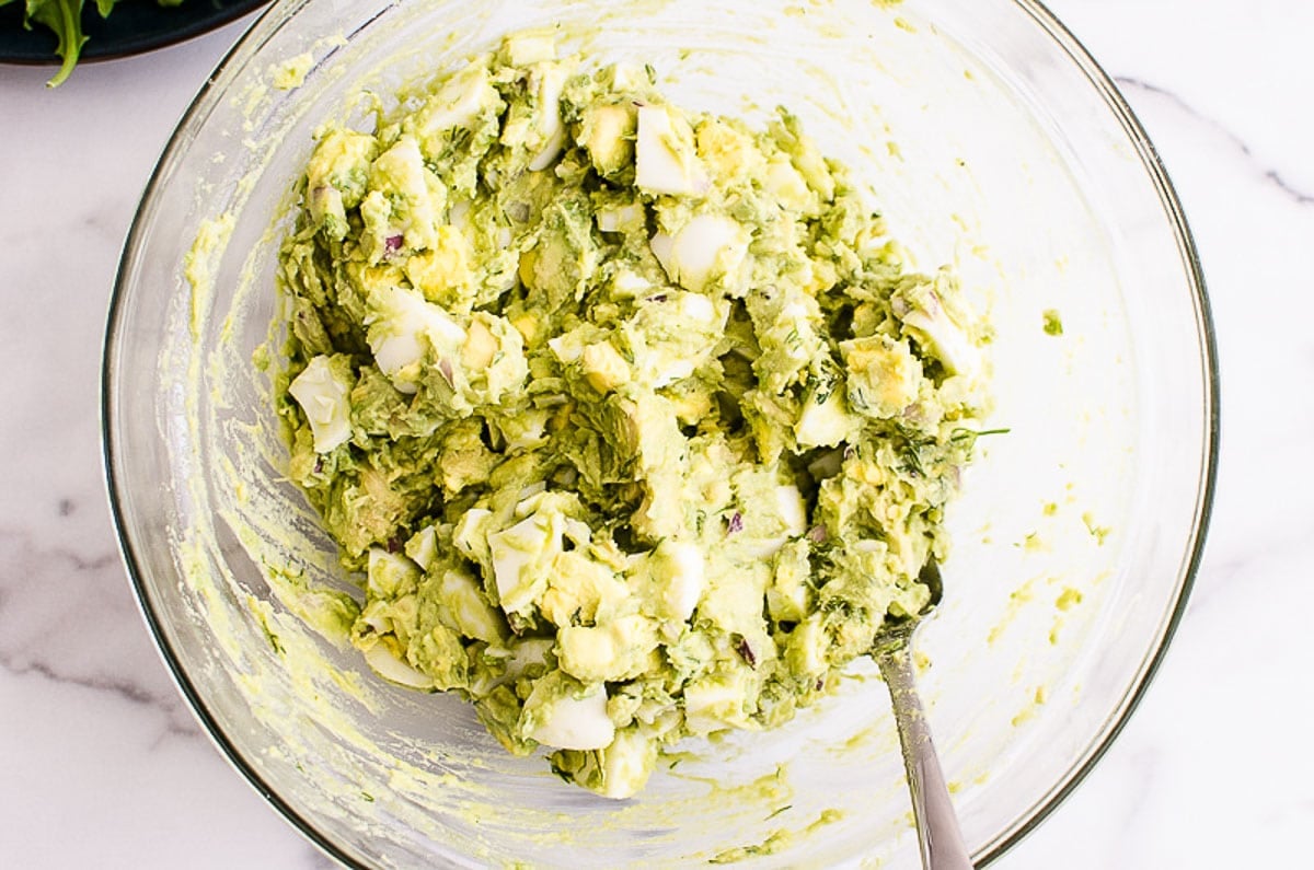 Mashed avocado with chopped egg in a bowl being mixed together with fork.