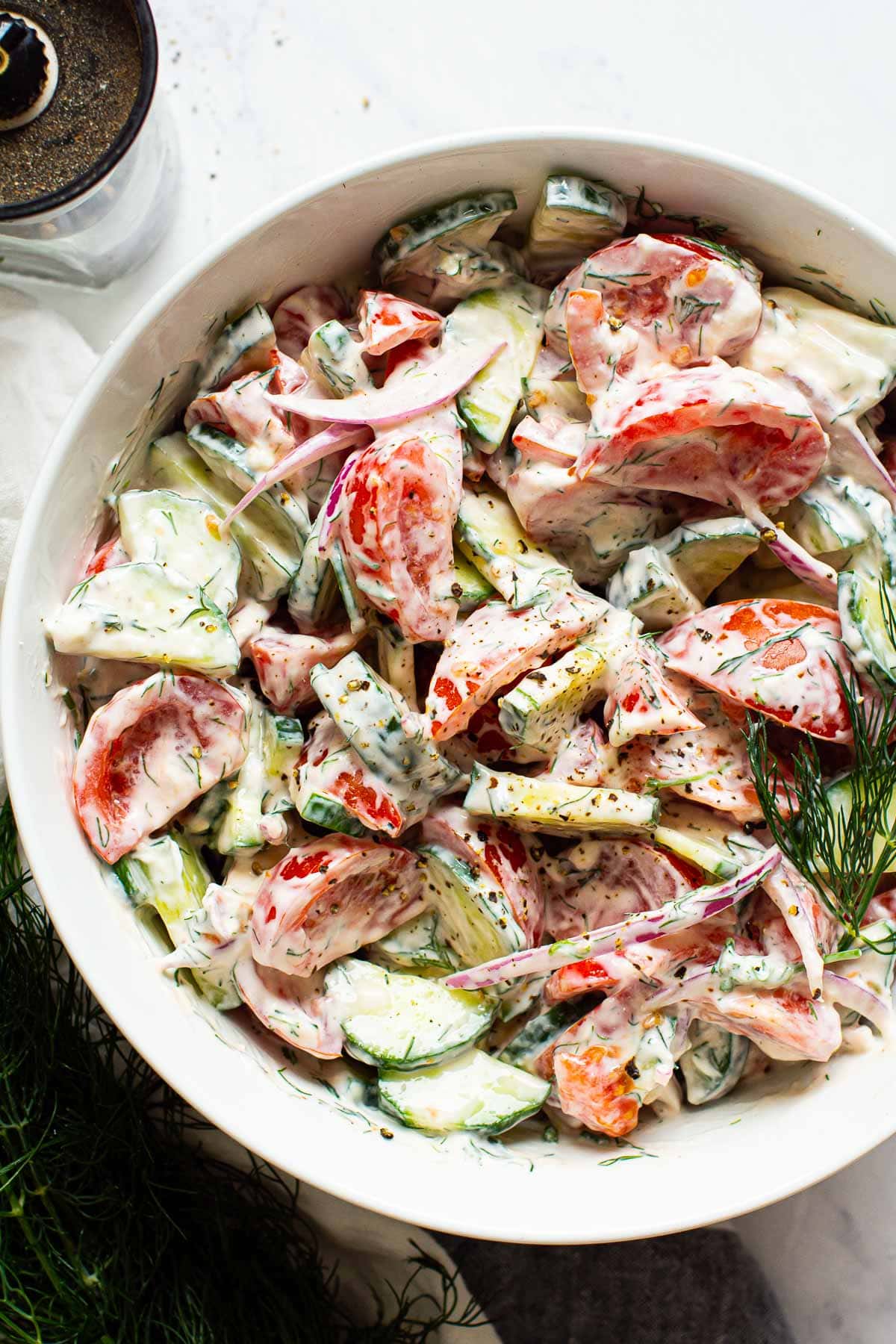 Creamy cucumber tomato salad in white bowl with pepper grinder and bunch of dill on a counter.