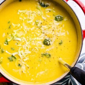 Healthy broccoli cheese soup garnished with cheese in red pot with ladle.