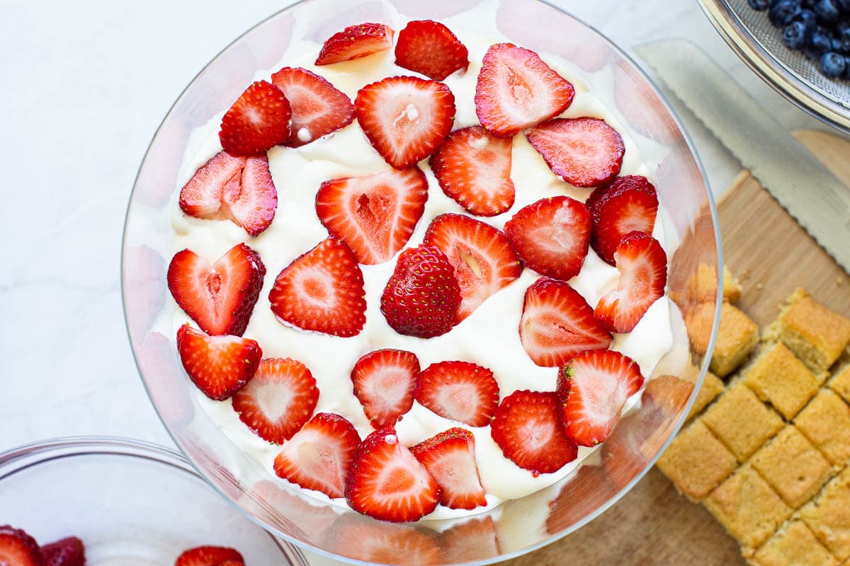 Strawberries sliced on top of cream in trifle dish. Cubed cake on cutting board.
