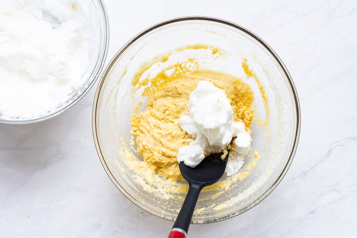 A scoop of egg whites on top of almond flour mixture in glass bowl with spatula.
