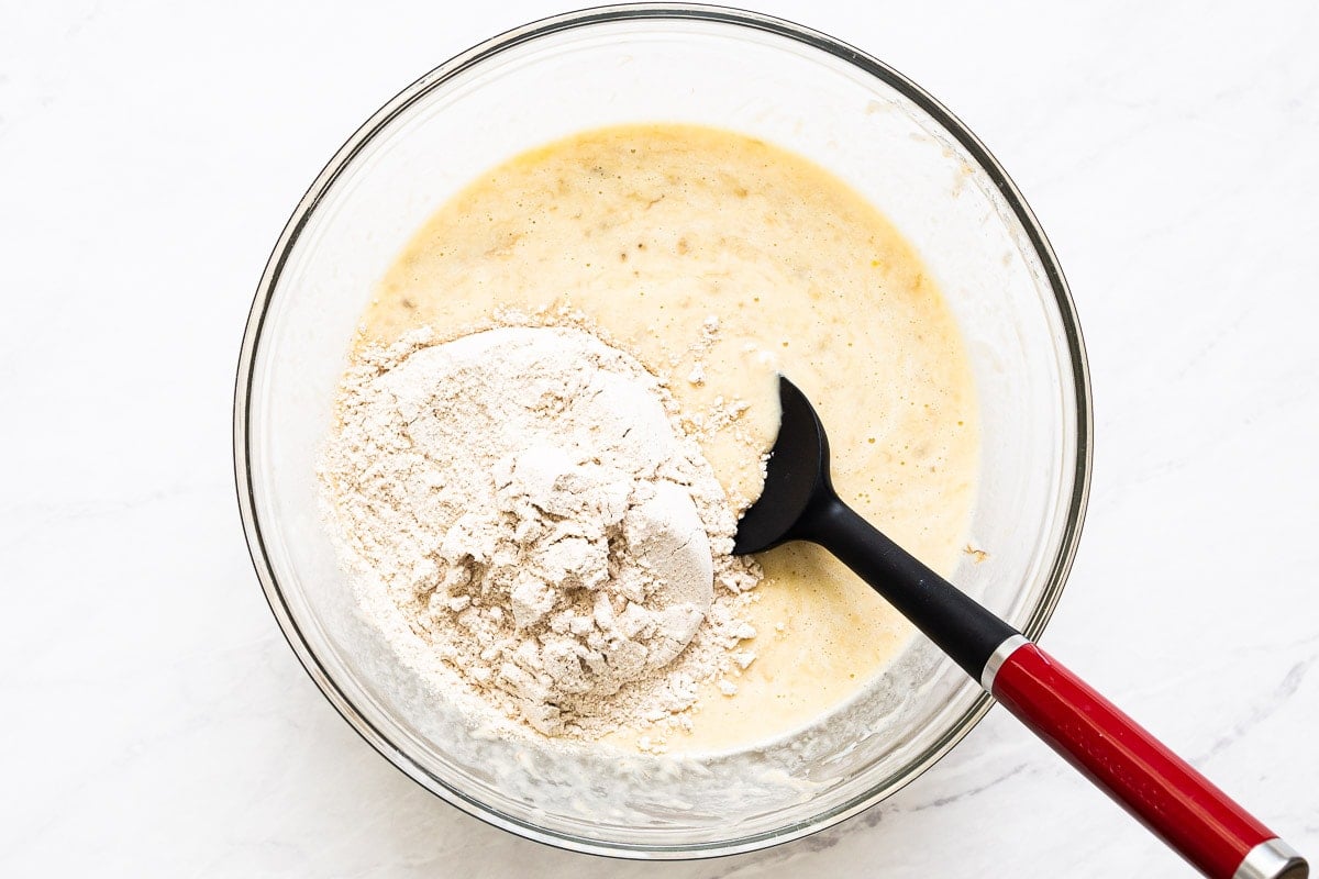 Flour being added to liquid mixture in bowl with spatula.