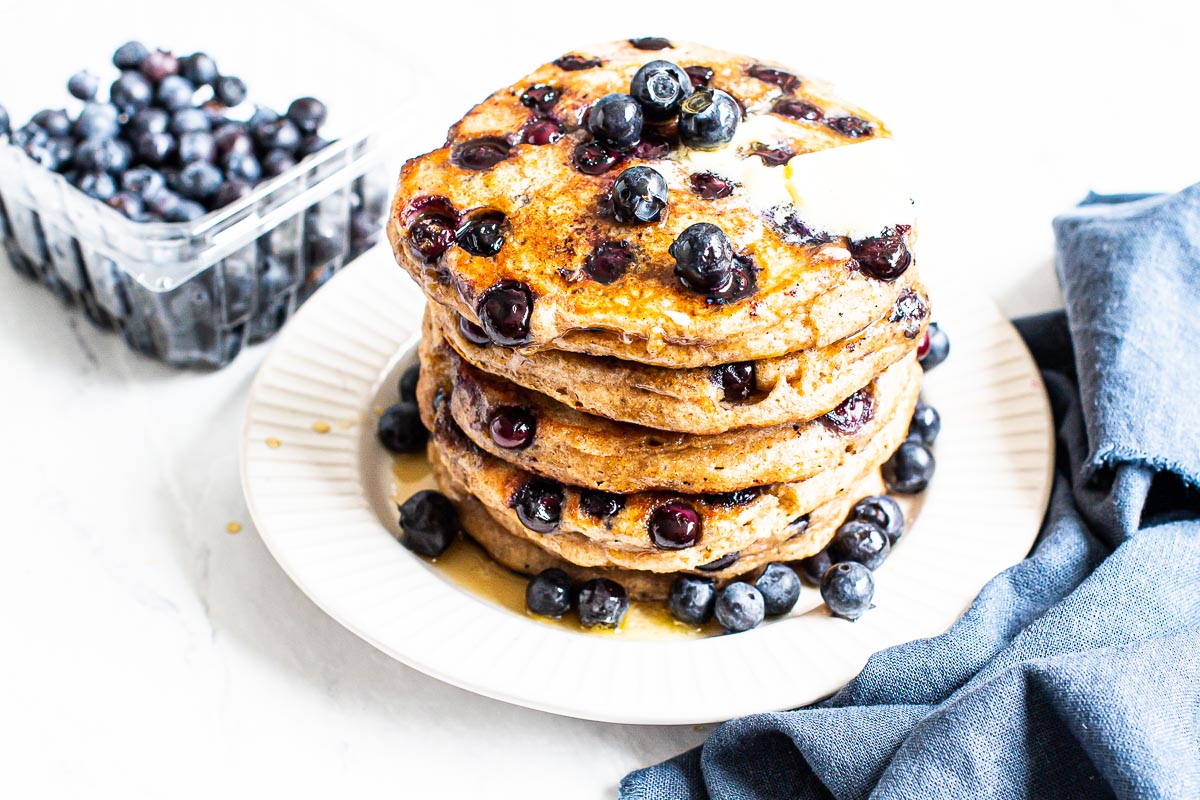 A stack of healthy blueberry pancakes with butter and container of fresh blueberries.