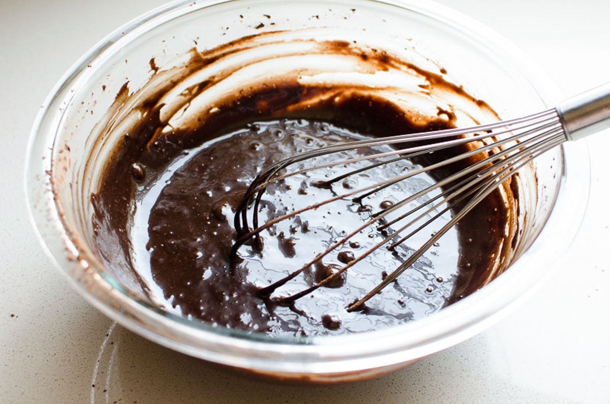 Liquid ingredients with cocoa powder in a bowl with whisk.