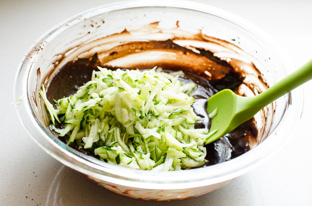 Shredded zucchini on top of liquid brownie batter with green spatula.