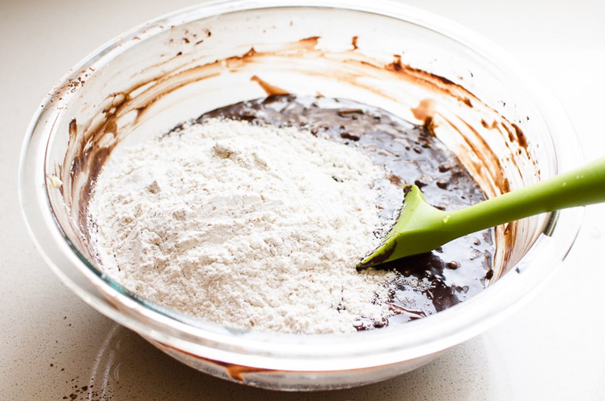Stirring flour into brownie batter with green spatula in glass bowl.