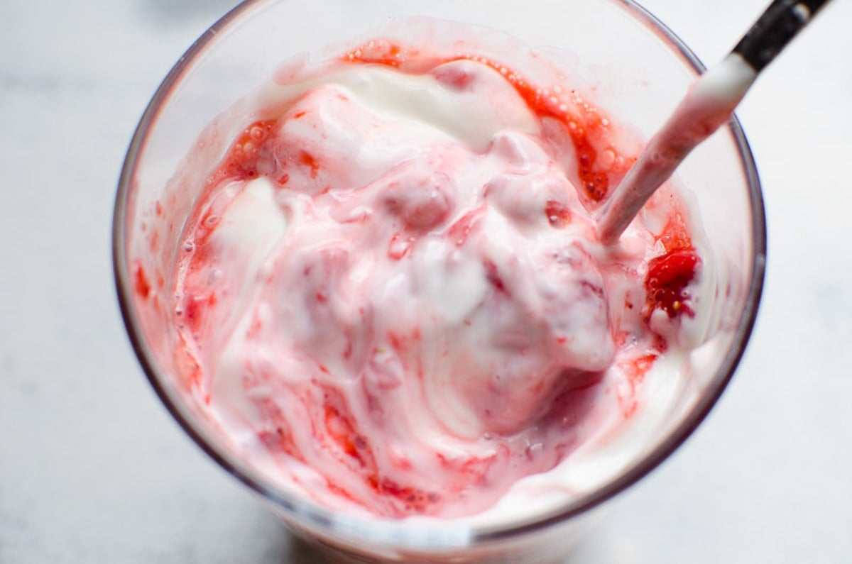 Yogurt and mushed strawberries stirred with a spoon in glass.