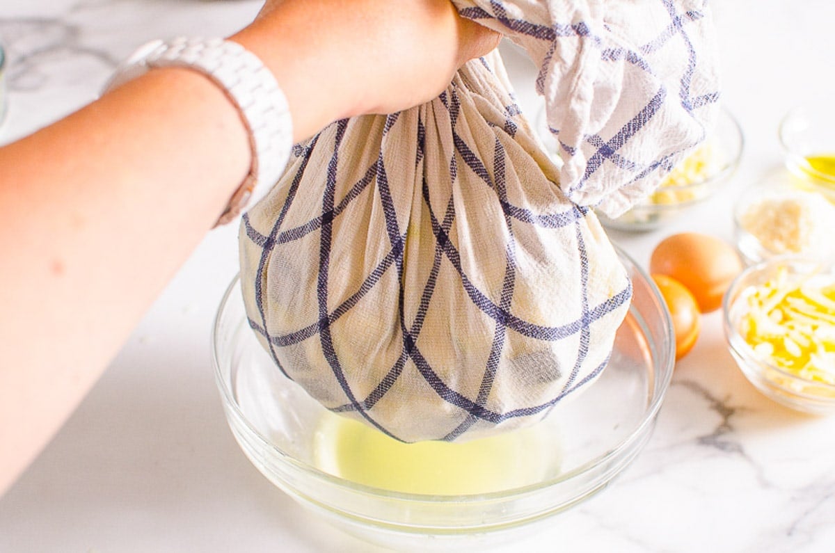 A hand holding zucchini in towel with a bowl containing excess zucchini water.