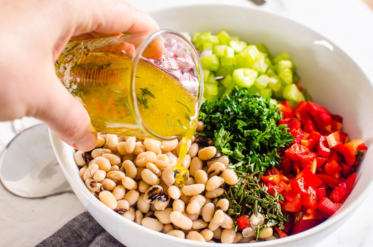 Pouring dressing over white bean salad ingredients in white bowl.