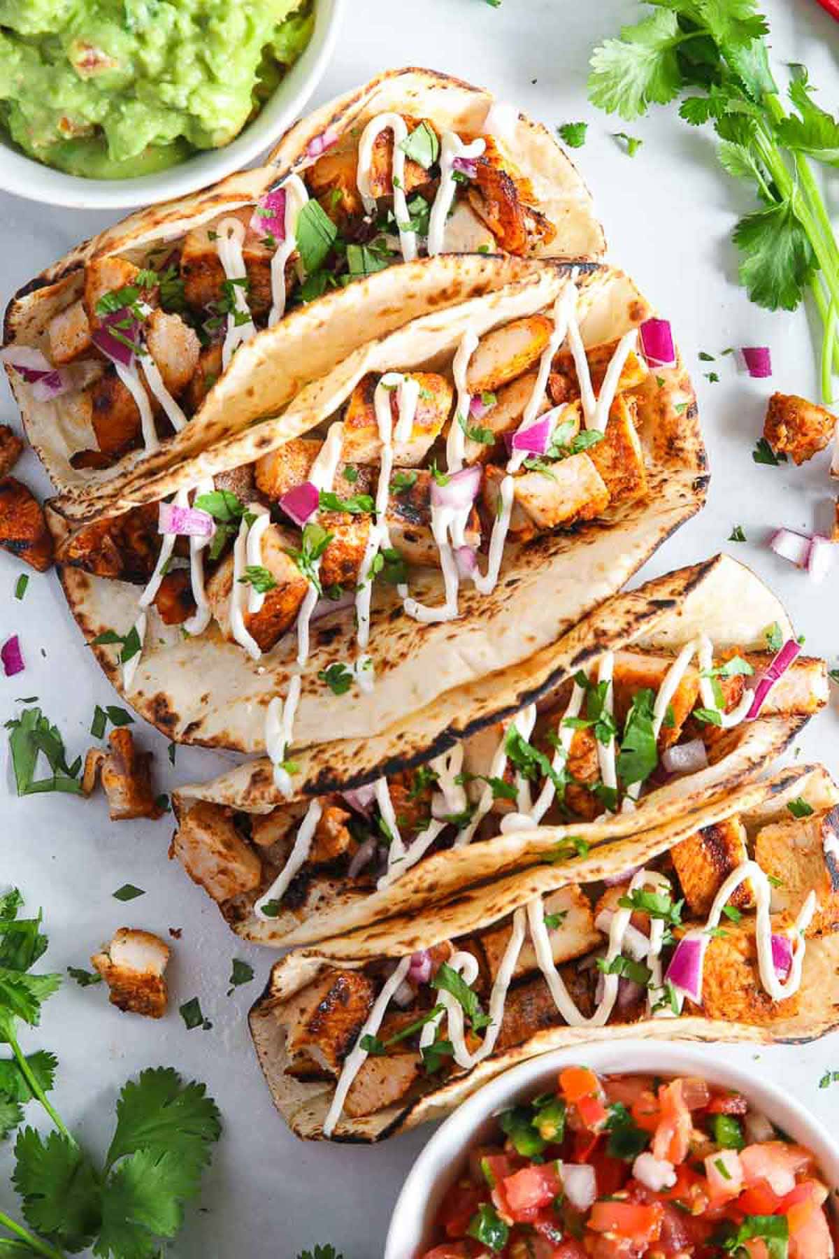 Grilled chicken street tacos on a plate garnished with cilantro, red onion and white sauce.