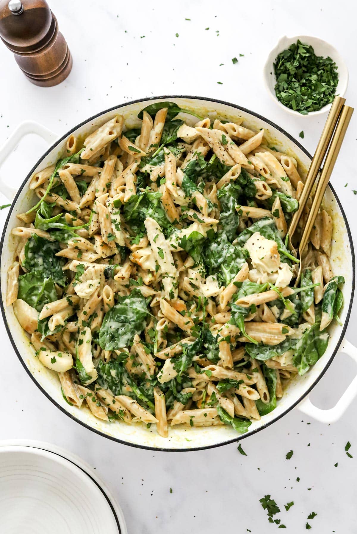 Chicken spinach pasta in a white pot with pepper grinder and small bowl of parsley.