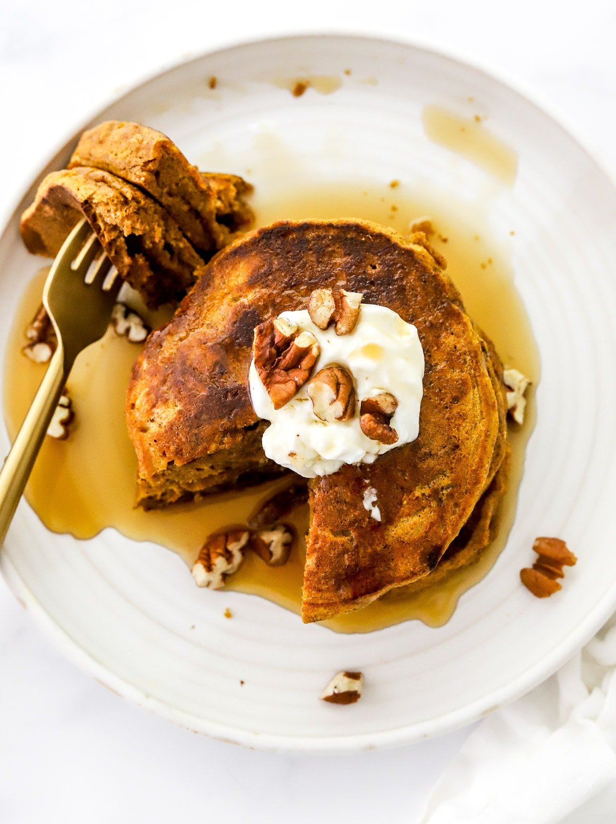 Looking down at a stack of healthy pumpkin pancakes with a bite on a fork and maple syrup, pecans and yogurt topping.