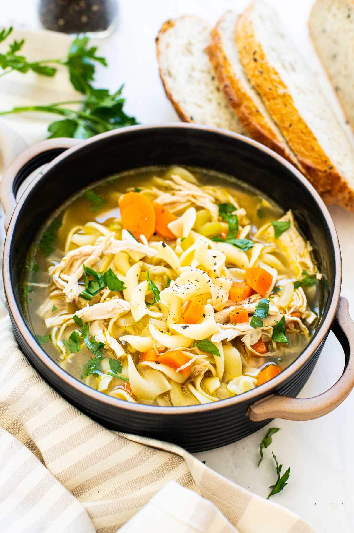 Turkey noodle soup in a bowl with parsley garnish. Sliced bread and linen on counter.