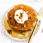 A stack of healthy pumpkin pancakes on a plate with maple syrup, pecans and yogurt.