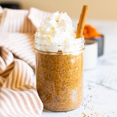 Pumpkin chia pudding in a jar topped with whipepd cream and cinnamon stick. Linen towel and spice on a counter.