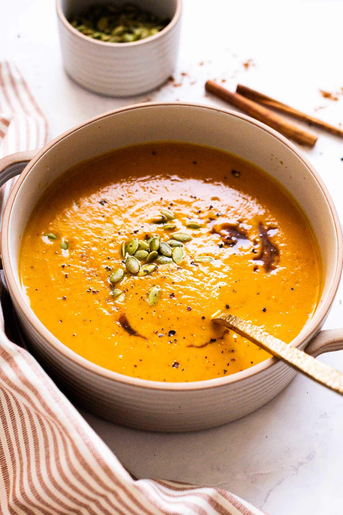 A bowl with healthy pumpkin soup with soy sauce, pumpkin seeds and spoon. Linen on a counter.