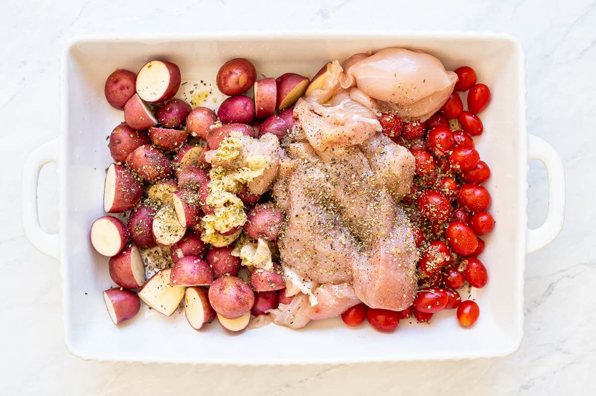 Chicken, tomatoes, potato halves, and seasonings in a white baking dish.