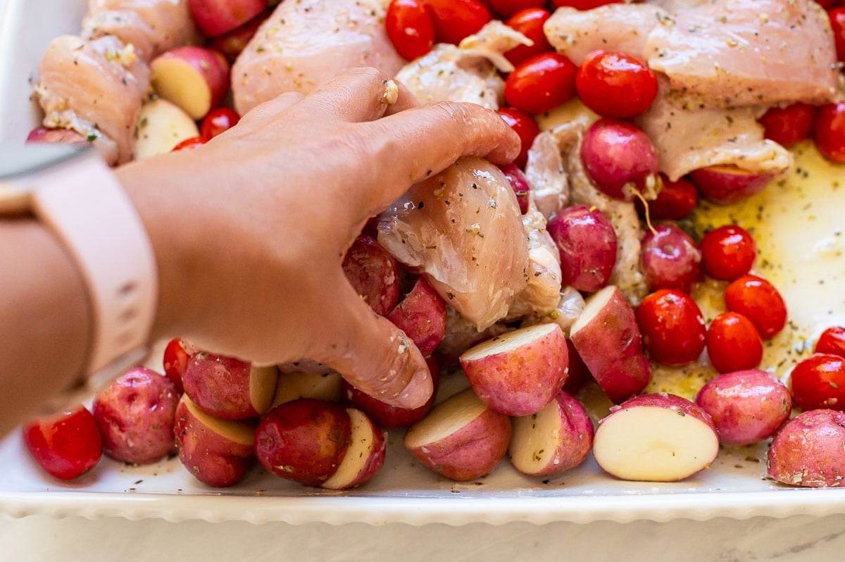 A hand mixing seasonings on chicken, tomatoes and potatoes together in baking dish.
