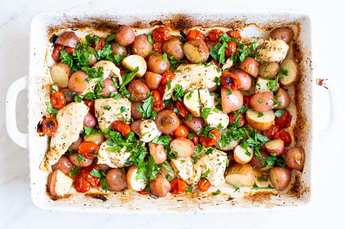 A white baking dish with cooked chicken, tomatoes, and potatoes garnished with parsley.
