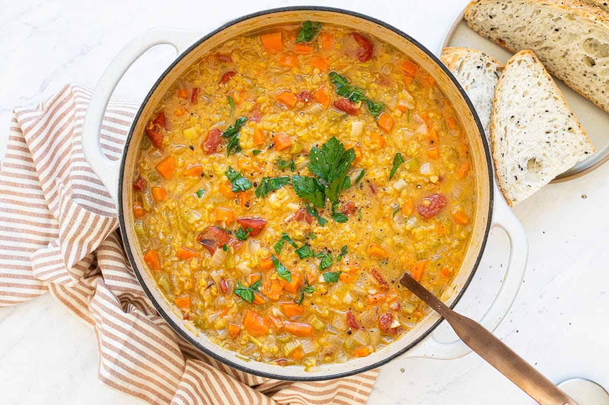 Red lentil vegetable soup garnished with parsley in white pot. Bread slices and linen towel on a counter.