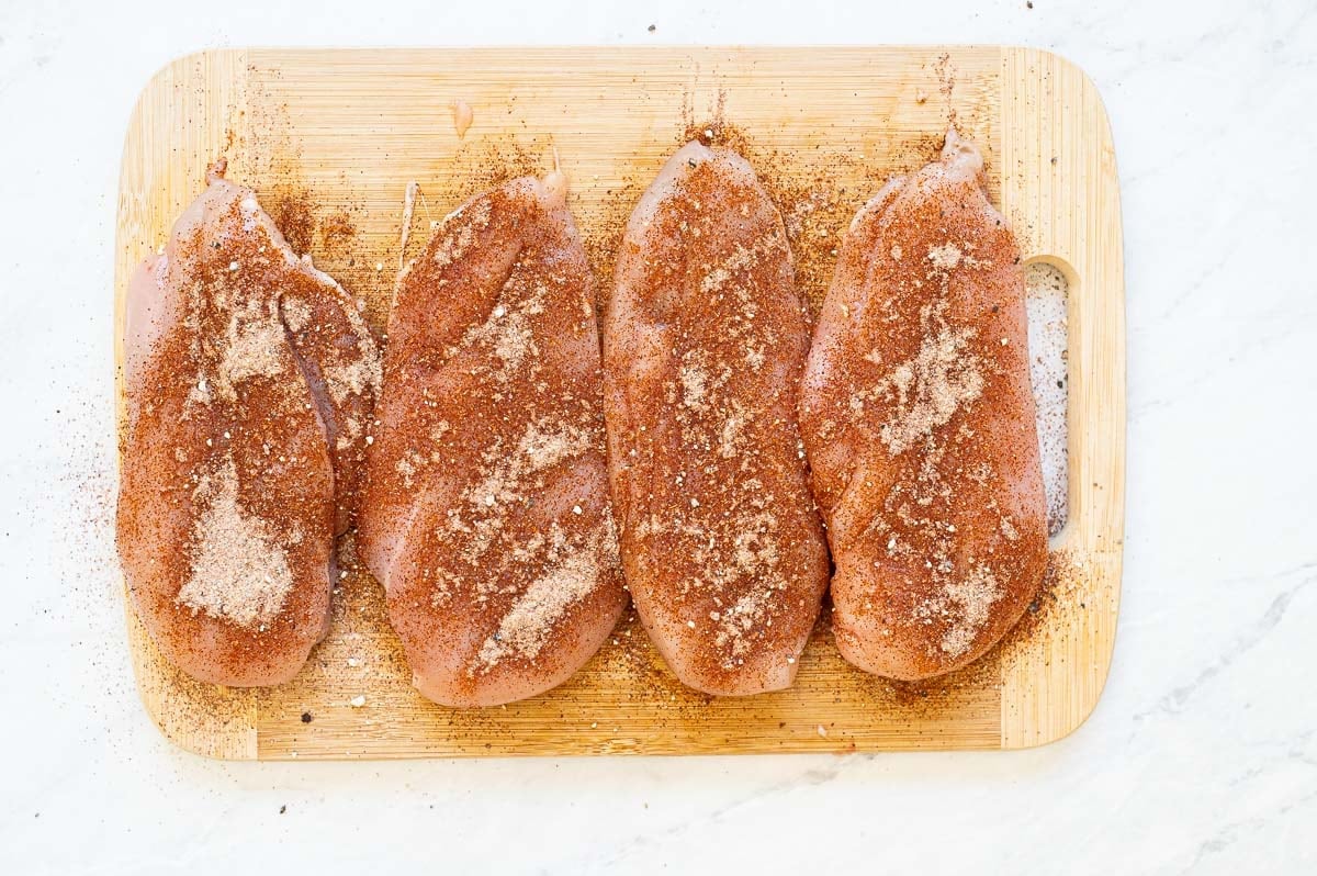 Four seasoned chicken breasts on a wooden board.