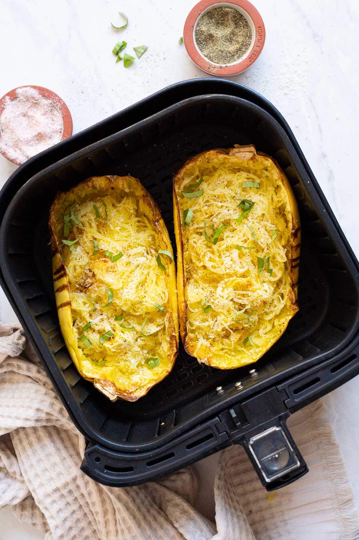 Two halves of spaghetti squash with cheese and basil in air fryer basket.