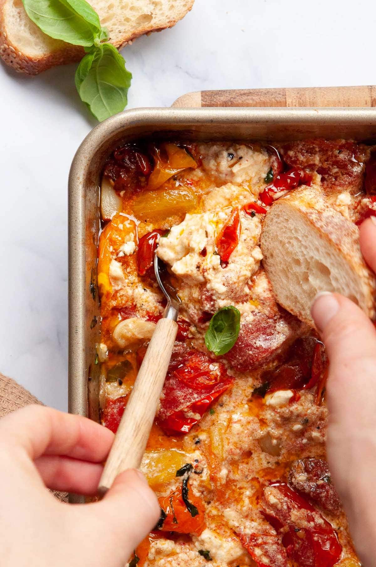 Person spooning some baked tomato feta dip on a slice of a baguette.