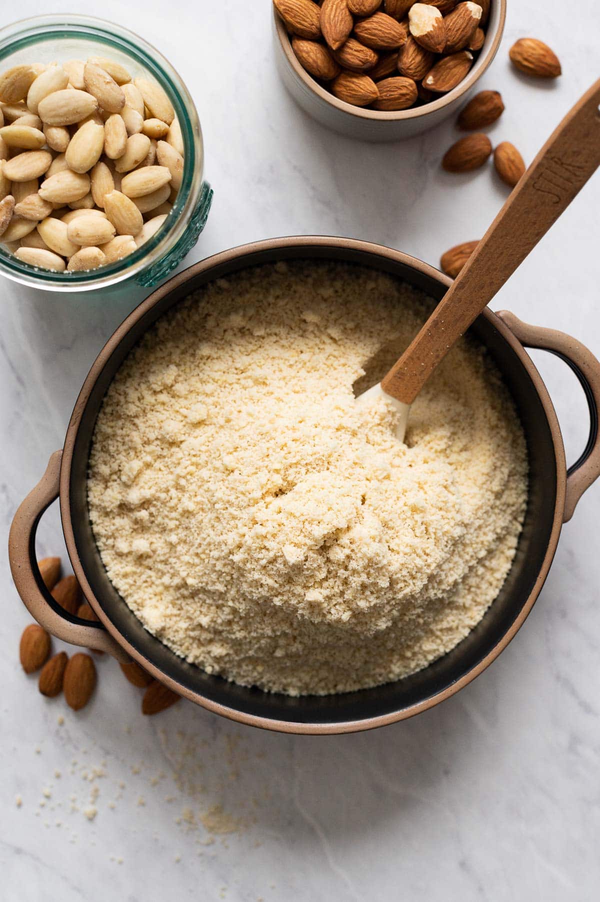 Homemade almond flour in black bowl with a spoon with blanched almonds around it.