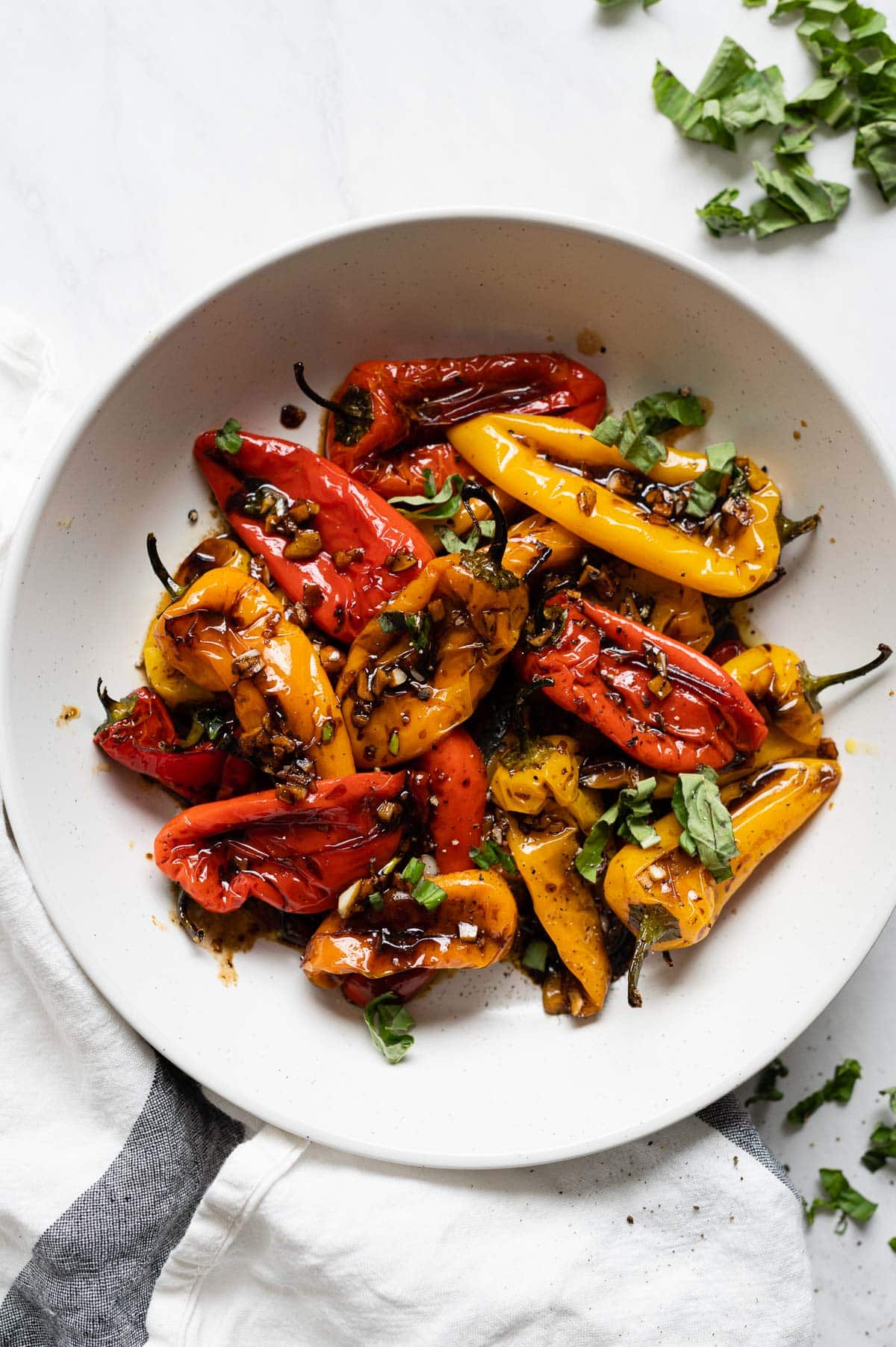 Yellow and red roasted mini sweet peppers garnished with basil and garlic in white bowl.