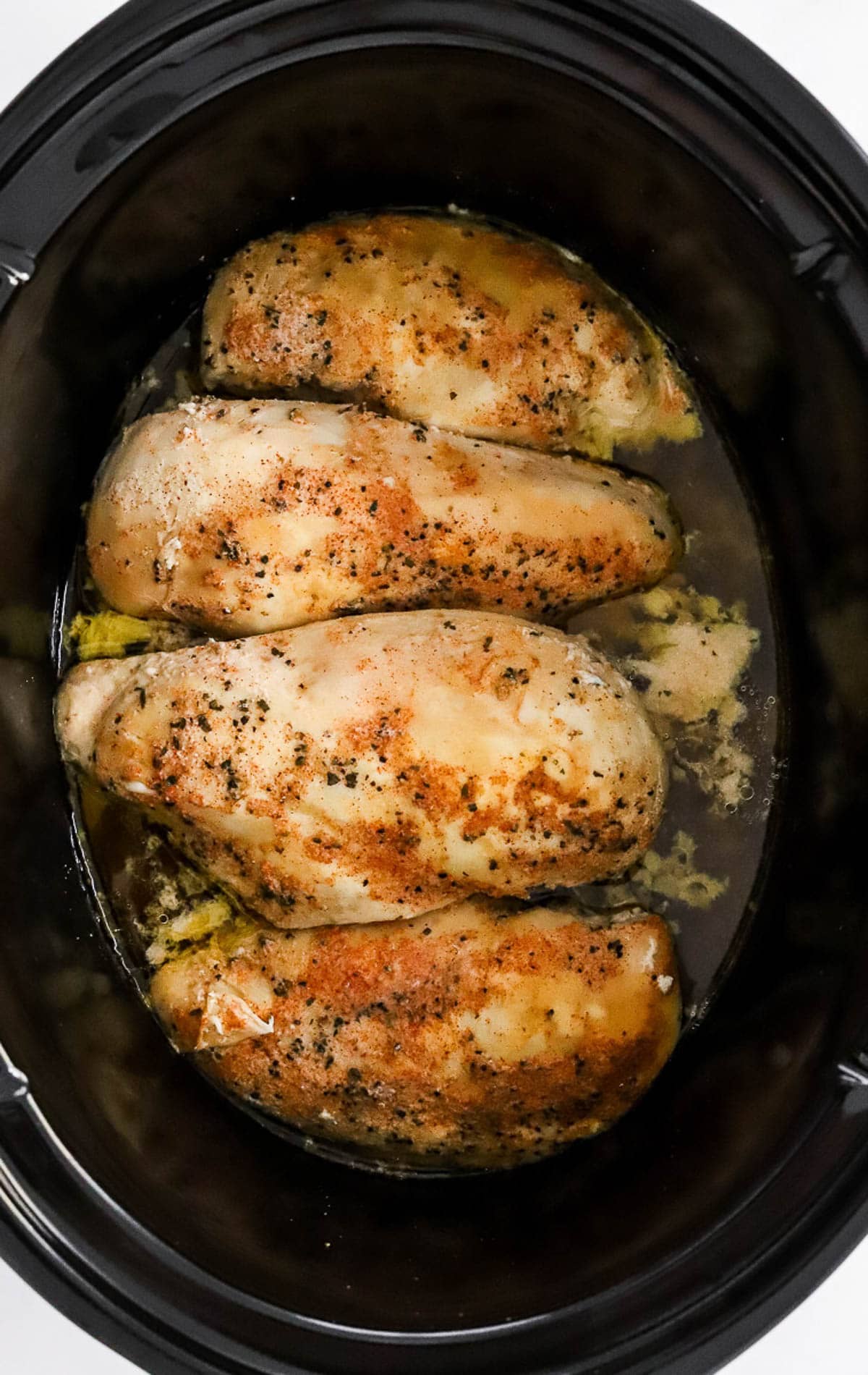 Four chicken breasts in black slow cooker with seasonings in broth.