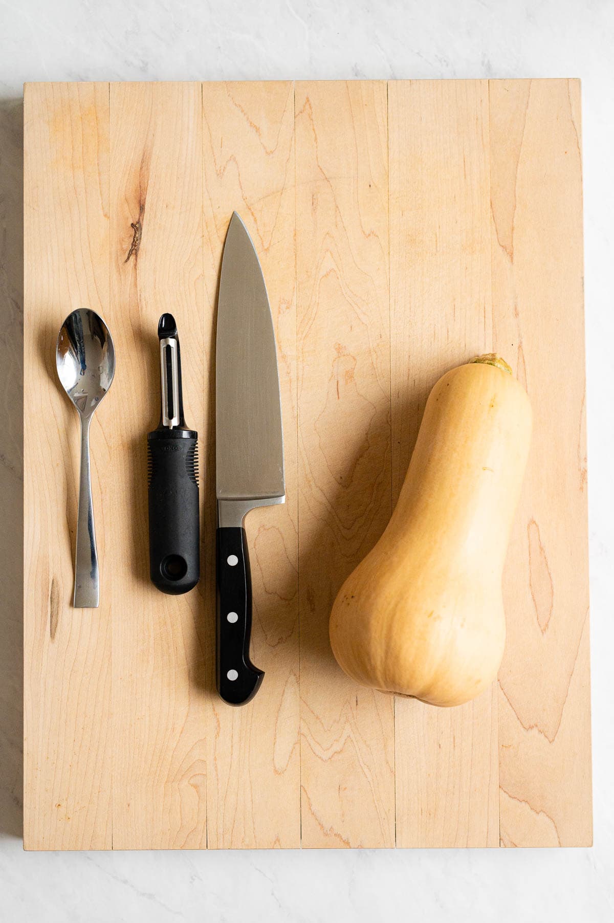 Butternut squash, chef's knife, vegetable peeler, tablespoon on large wooden cutting board.