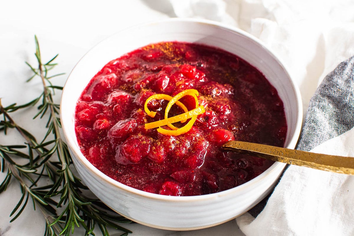 Fresh cranberry sauce in white bowl with golden spoon and orange rind on top.