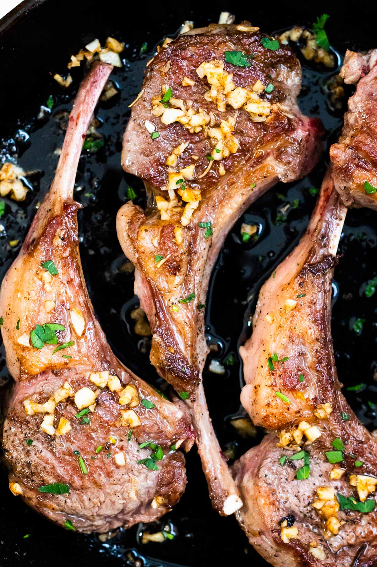 Close up of three pan-seared lamb chops with garlic butter and fresh parsley in cast iron skillet.