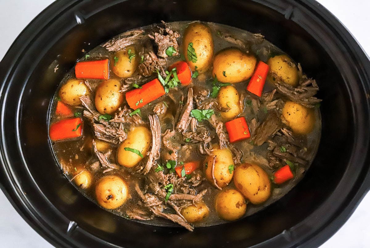 Cooked shredded roast with baby potatoes and carrots in gravy in black slow cooker.