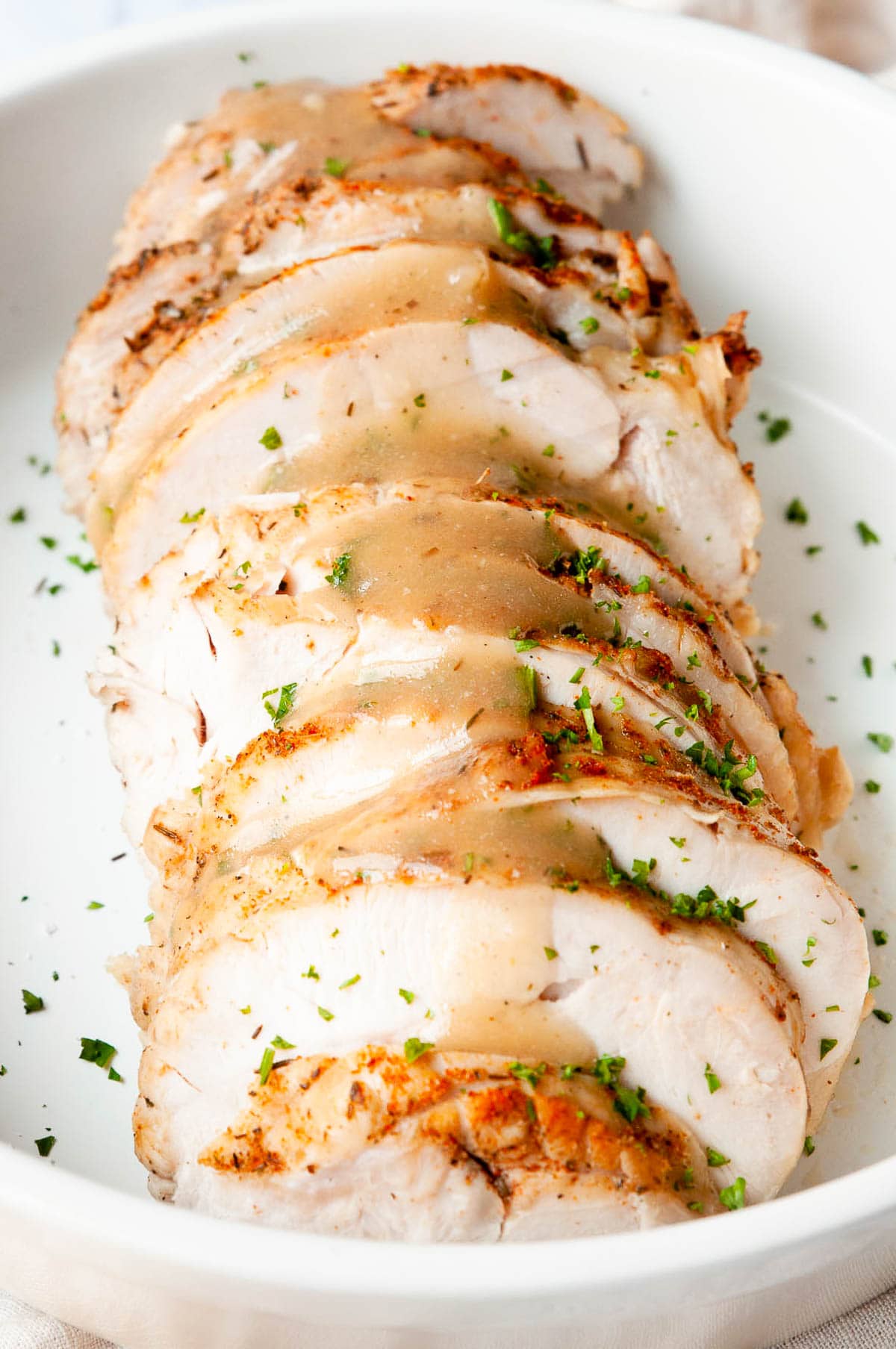 Cooked and sliced boneless turkey breast with gravy and parsley on white plate.