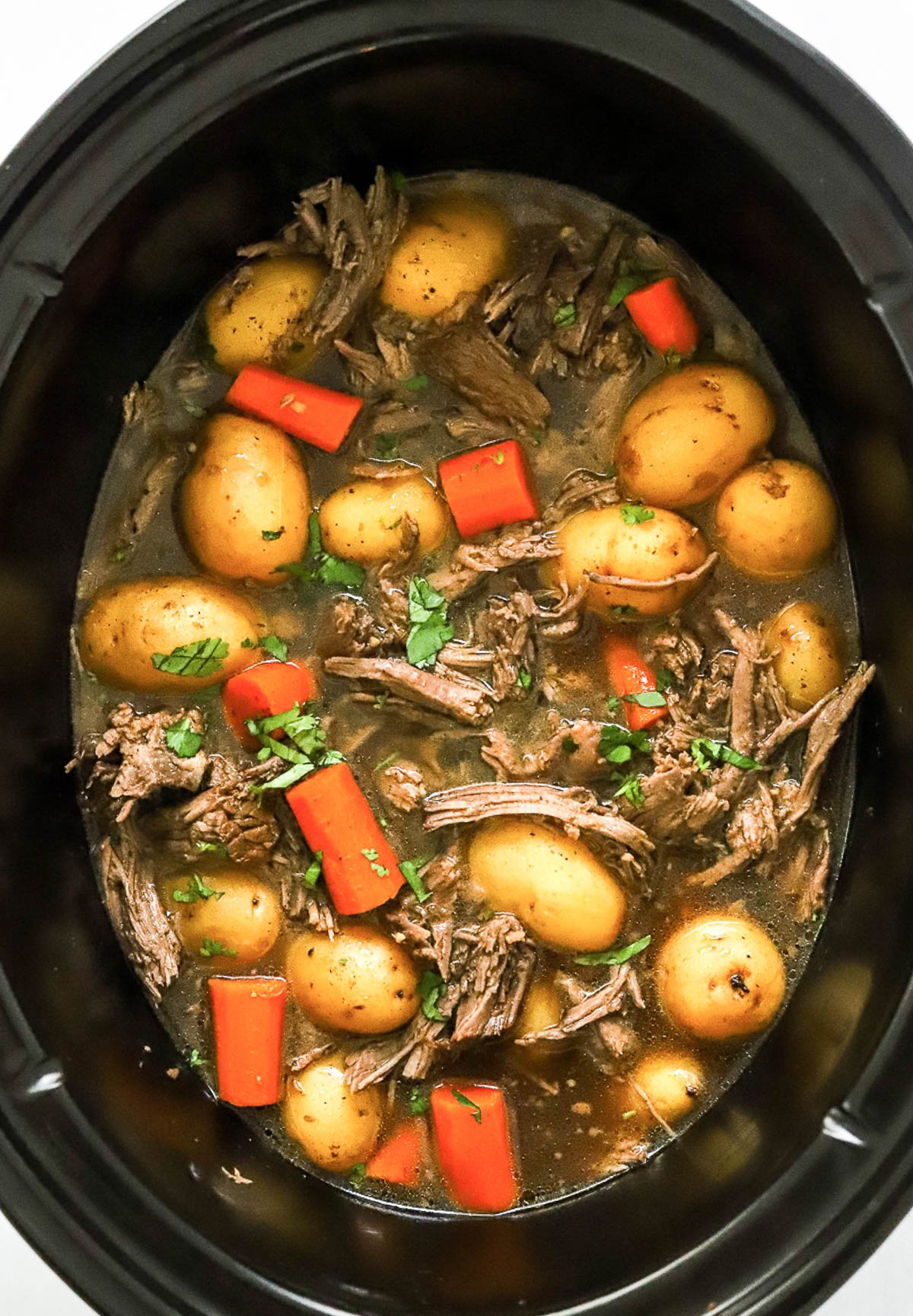 Shredded rump roast in crock pot with baby potatoes and carrots in broth and garnished with parsley.