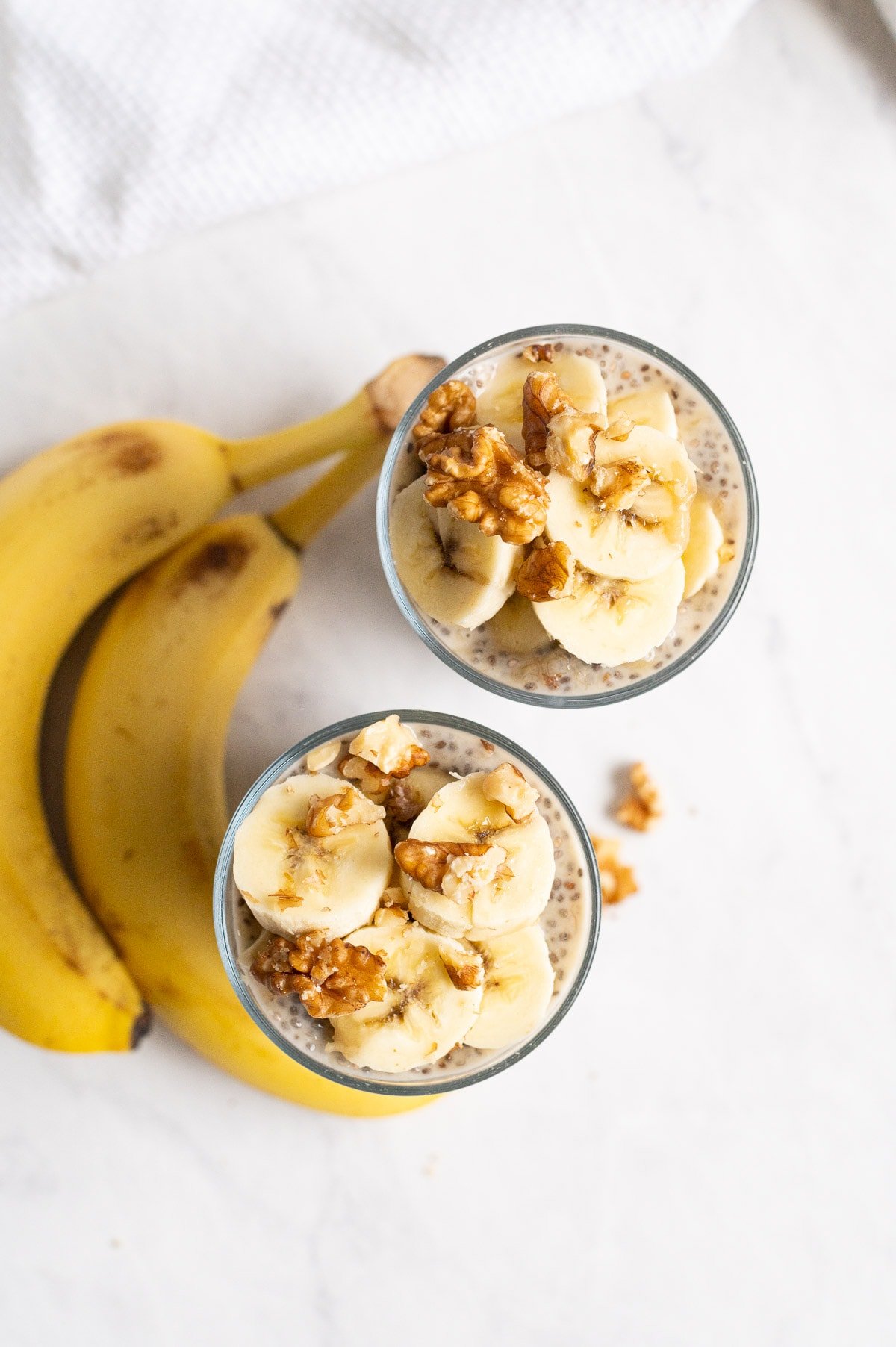 Two glasses with banana chia pudding topped with sliced bananas, walnuts and whole bananas on a countertop.