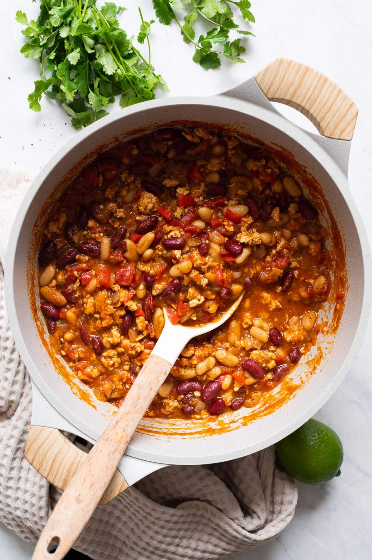 Ground chicken chili in white pot with a spoon. Lime, cilantro and linen towel on a counter.