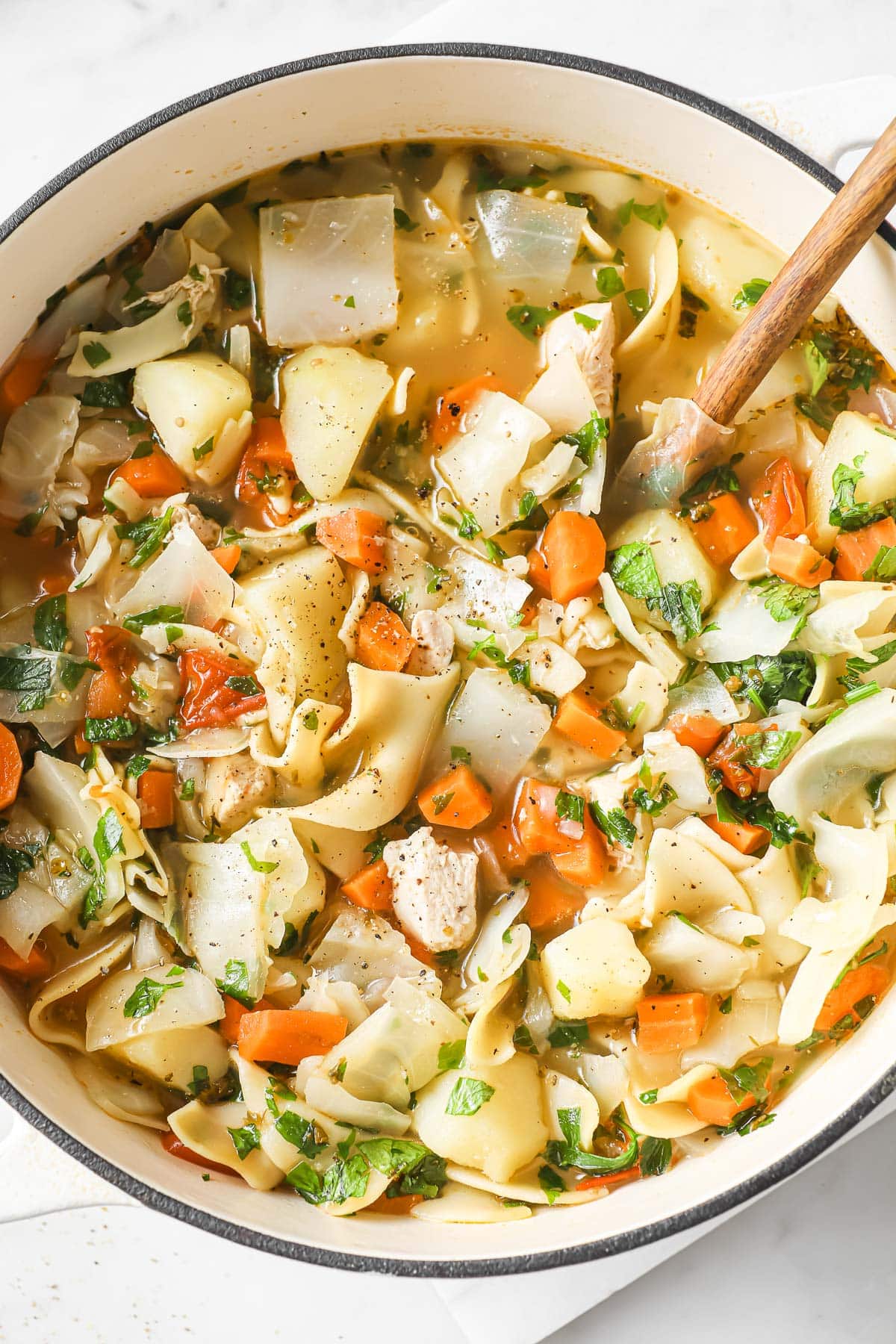 White pot of chicken noodle vegetable soup and wooden spoon in it.