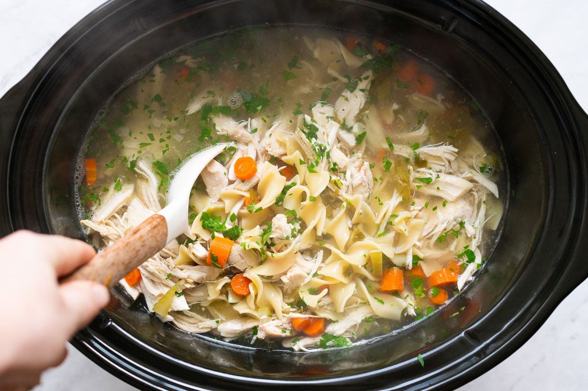 Person steering slow cooker chicken noodle soup with a spoon.