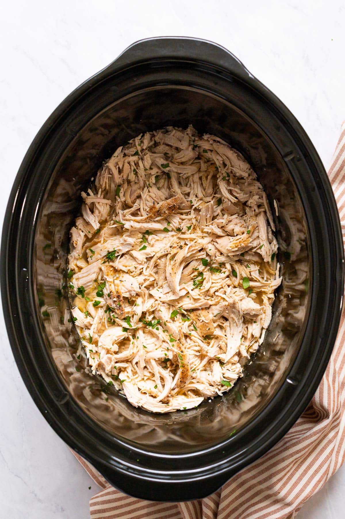 Shredded chicken in broth garnished with herbs in black slow cooker.