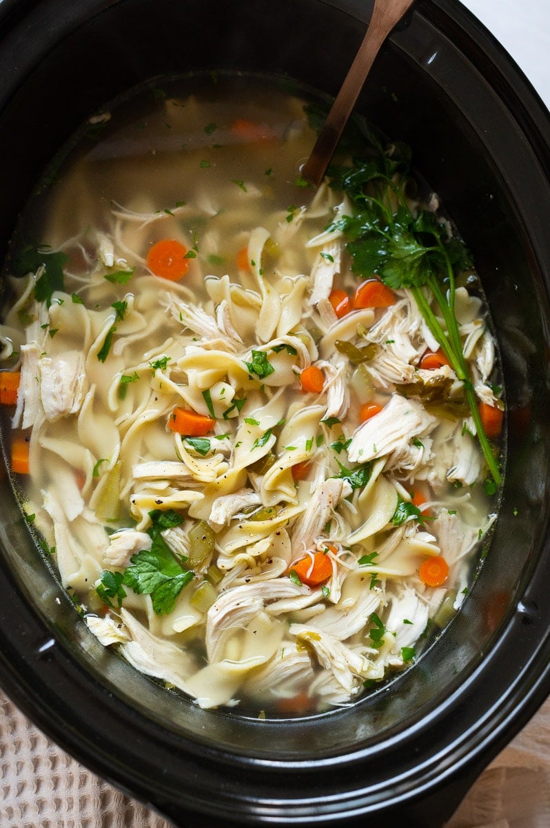 Crockpot chicken noodle soup with parsley in black slow cooker.