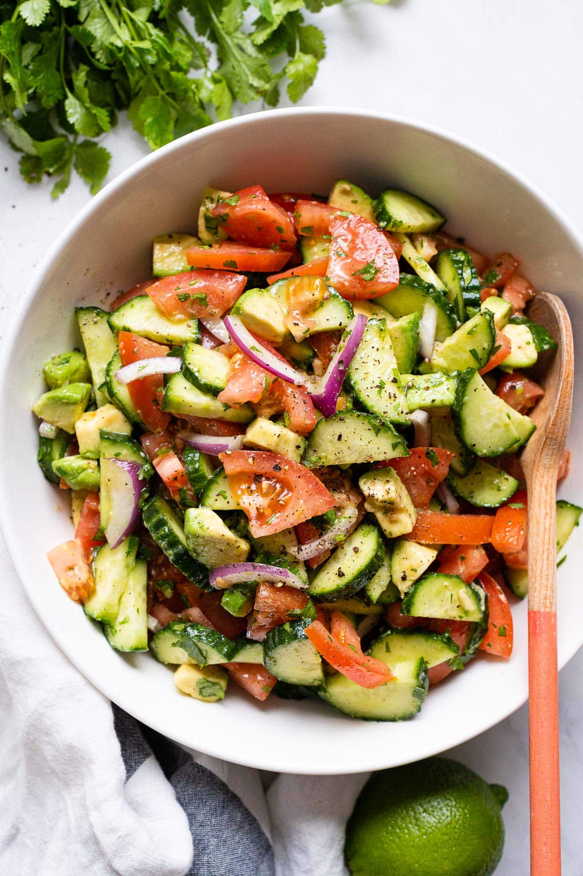 Cucumber tomato avocado salad in white bowl with wooden spoon.